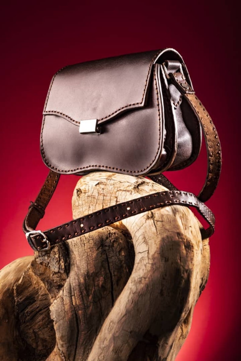 May include: A brown leather satchel bag with a silver buckle and a long strap. The bag is sitting on a piece of weathered wood.