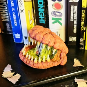 May include: A unique pencil sharpener shaped like a monster's mouth, with sharp, yellow teeth and a textured orange exterior. The interior is green, and a pencil is inserted for sharpening. Pencil shavings and a pencil are on the black surface.