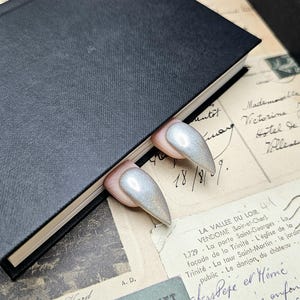 Puede incluir: Dos diseños de uñas largos con purpurina plateada y base rosa claro se muestran junto a un libro negro cerrado. Las uñas tienen forma de almendra y un acabado brillante e iridiscente. En el fondo, objetos efímeros vintage, como postales y entradas.