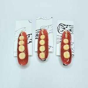 Könnte beinhalten: Drei rote, längliche Dekorationsartikel, jeweils mit vier cremefarbenen, kreisförmigen Akzenten. Sie sind auf kleinen weißen Karten mit einem schwarz-weißen Design befestigt. Die Artikel haben eine glänzende Oberfläche.