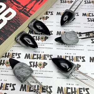 Puede incluir: Una colección de clips de papel únicos con detalles esculpidos en negro y gris. Los clips de papel tienen un acabado brillante y se muestran sobre una superficie estampada con el texto "THIS IS MICHIE'S SHOP".