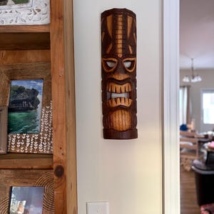 Puede incluir: Una máscara tiki de madera tallada con un diseño detallado. La máscara es marrón con detalles de madera más claros, con una expresión feroz con la boca abierta y dientes visibles. Está montada en una pared de color crema.