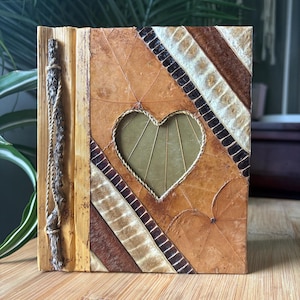 May include: A rectangular journal with a cover made of layered brown and tan dried leaves. The cover features a central heart-shaped cutout, diagonal striped patterns of dark brown and cream, and a spine decorated with a thin, textured twig and twine.