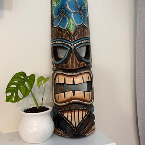 Puede incluir: Una máscara tiki de madera tallada, principalmente marrón con detalles florales azules. La máscara presenta aberturas para los ojos y la boca. Se exhibe en una estantería blanca, junto a una planta en maceta. Objeto decorativo.