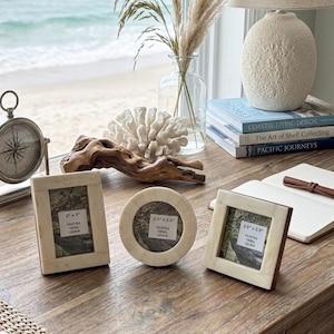 May include: Three decorative photo frames with off-white and brown accents are displayed on a wooden desk. The frames are rectangular and circular, with the text "PASSTIKA TIDAL LOERIA" and "PALESTINA TRIBAL LOCATR" visible. A compass, vase, and books are also on the desk.