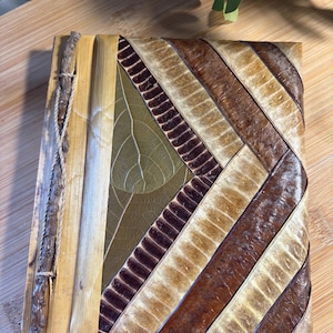 May include: A rectangular journal with a cover crafted from layered, dried plant leaves in shades of brown, tan, and olive green. The cover features a chevron pattern and is bound with a thin, natural fiber cord wrapped around a small twig.