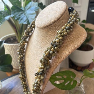 May include: A handcrafted necklace composed of small, irregularly shaped, light green and tan beads. The necklace is displayed on a tan bust form, showcasing its intricate design and natural color palette. The clasp is a dark, rounded bead.
