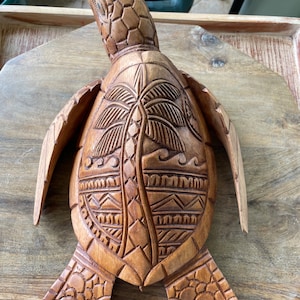 Pode incluir: Escultura de tartaruga de madeira com entalhes intrincados. A carapaça da tartaruga apresenta um desenho de palmeira e padrões geométricos. A escultura é de cor castanha quente e parece ser feita à mão.
