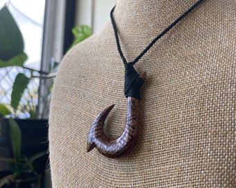 Puede incluir: Un collar con colgante de gancho de madera marrón con un cordón negro. El gancho tiene intrincados patrones tallados y se exhibe en un busto cubierto de arpillera. El collar es una pieza de joyería artesanal.