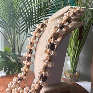 May include: A handcrafted necklace featuring a repeating pattern of light brown and dark brown beads, interspersed with white seashells. The necklace is displayed on a tan jewelry bust against a backdrop of green plants and a wooden table.