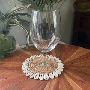 May include: A clear glass goblet sits atop a circular woven coaster. The coaster features a natural tan rattan centre surrounded by a decorative border of white cowrie shells arranged in a radial pattern around the outer edge of the base.