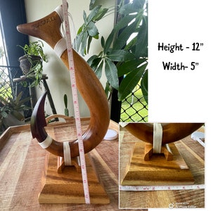 May include: A wooden Hawaiian-style fish hook sculpture. The sculpture is 12 inches tall and 5 inches wide. It features a curved design with a dark brown hook and a lighter brown body, with the word "HAWAII" carved into the top.