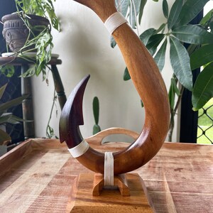 May include: A wooden fish hook sculpture with the word "HAWAII" carved into the top. The hook is brown with a dark brown tip and white rope accents. It is mounted on a square wooden base.