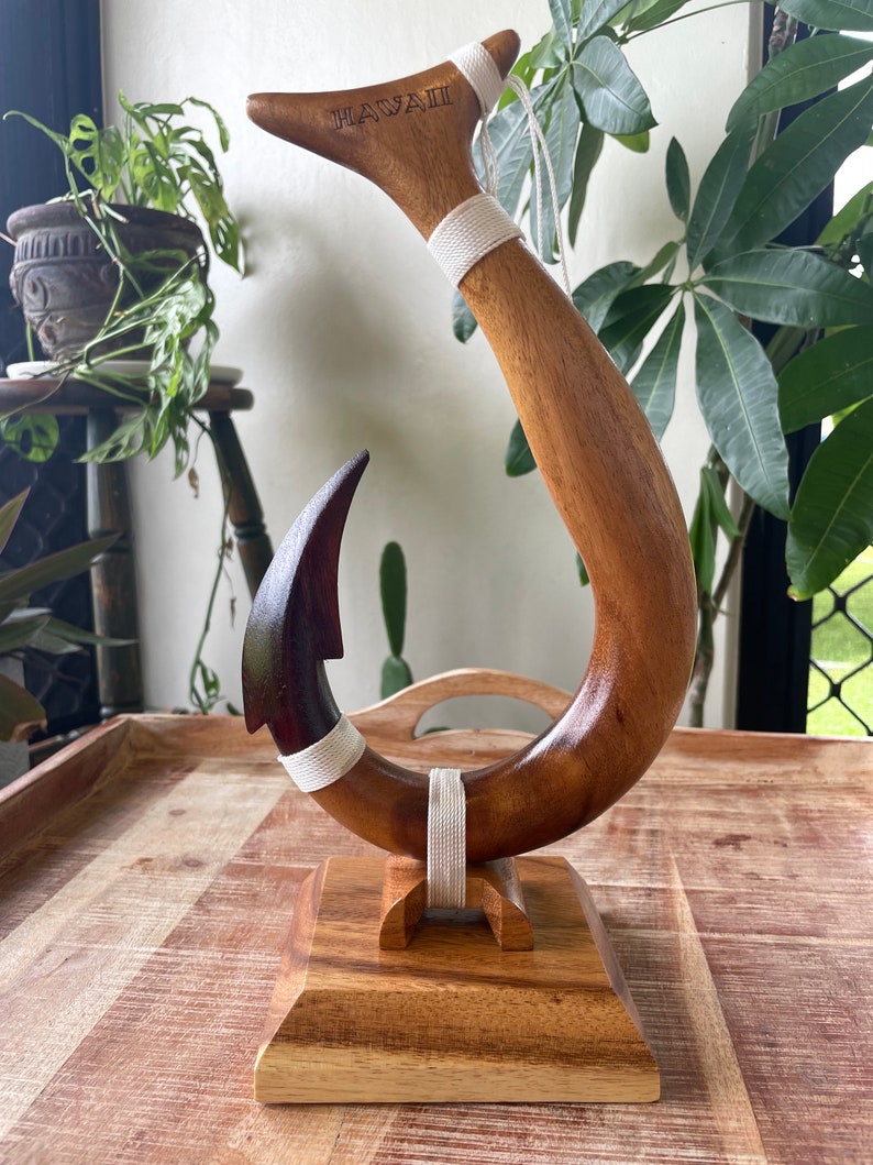 May include: A wooden fish hook sculpture with the word "HAWAII" carved into the top. The hook is brown with a dark brown tip and white rope accents. It is mounted on a square wooden base.