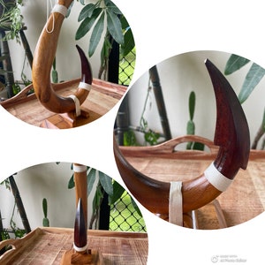 May include: A handcrafted wooden sculpture with a curved, horn-like shape. The sculpture features a dark brown tip and a lighter brown body, accented with a white fabric band. It is mounted on a small wooden base.