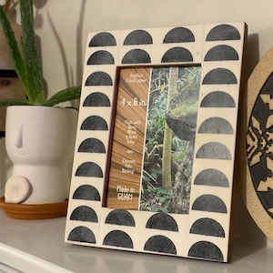 May include: A decorative photo frame with a cream-colored border featuring a repeating black half-circle design. The frame holds a 4 x 6 in. photo and is made with wood, resin inlay, and coconut palm backing. The text "Made in GUAM" is visible.