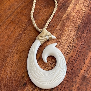 Op de afbeelding: Crèmekleurige hangerketting met een gesneden, gebogen haakontwerp. De haak is versierd met geëtste patronen en hangt aan een gevlochten beige koord. De ketting wordt weergegeven op een houten oppervlak.