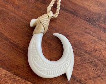 May include: Off-white pendant necklace featuring a carved bone hook design. The hook is intricately detailed with a leaf-like pattern and is suspended from a braided beige cord. The hook is approximately 3 inches long.