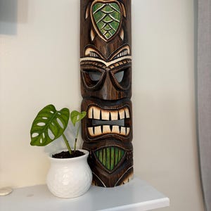 Puede incluir: Una máscara tiki de madera tallada con un diseño de tortuga y una boca abierta sonriente. La máscara es marrón con acentos verdes y blancos. Una pequeña planta en maceta en una maceta blanca texturizada está al lado de la máscara.