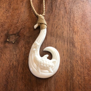May include: Cream-colored carved bone pendant necklace featuring a turtle design within a wave. The pendant is suspended from a tan braided cord, with the top portion wrapped in a matching twine. The necklace rests on a dark wood surface.