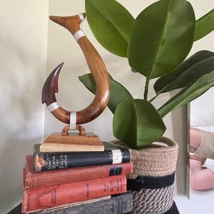 May include: A wooden fish hook sculpture with a dark red hook and light brown handle, resting on a wooden base. The sculpture is placed on a stack of vintage books. A green plant in a woven basket is in the background.