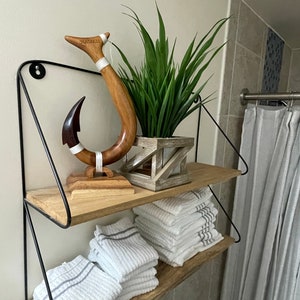 May include: A two-tiered shelf with a black metal frame. The top shelf holds a wooden hook sculpture with the word "HAWAII" and a plant in a geometric wooden planter. The bottom shelf has folded white towels.