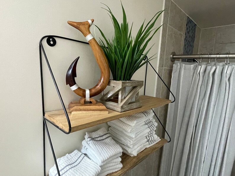 May include: A two-tiered shelf with a black metal frame. The top shelf holds a wooden hook sculpture with the word "HAWAII" and a plant in a geometric wooden planter. The bottom shelf has folded white towels.