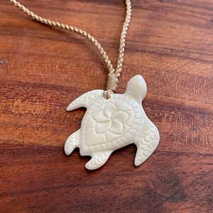 May include: Cream-colored turtle pendant necklace with a braided tan cord. The turtle pendant features a carved floral design on its shell. The necklace is displayed on a wooden surface.