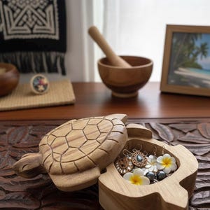 Peut inclure: Un coffret à bijoux en forme de tortue en bois, ouvert pour révéler des boucles d'oreilles et des fleurs. La boîte est marron clair avec des détails sculptés sur le couvercle. La boîte est sur une surface en bois sculpté brun foncé.