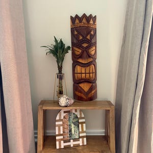 Puede incluir: Una estatua tiki de madera tallada con una corona se encuentra en una estantería de madera, junto con un marco de fotos, una concha y una planta en un jarrón dorado. El tiki es marrón con detalles tallados. El marco de la foto tiene un diseño geométrico blanco y marrón.