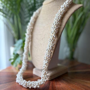 May include: A long, white beaded necklace draped over a mannequin. The necklace is made of small, round beads, creating a textured, rope-like appearance. The background includes greenery and a wooden surface.