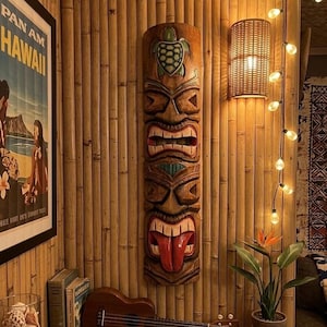 Puede incluir: Una máscara tiki de madera tallada con un diseño de tortuga y una lengua roja cuelga de una pared de bambú. También se ve un póster enmarcado de "Pan Am Hawaii", un ukelele y una planta en maceta con flores naranjas.