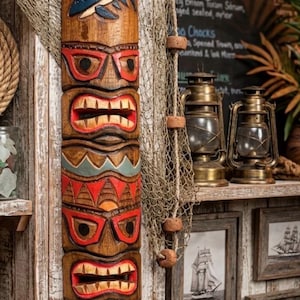 Puede incluir: Una estatua tiki de madera tallada con un diseño de tiburón, detalles rojos y naranjas, y una boca sonriente. Se exhibe en una pared de madera rústica con una red de pesca y grabados náuticos enmarcados. El texto "ISLAND COCKTAIL" es visible.