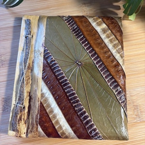 May include: A rectangular journal with a cover made of layered, dried botanical leaves in shades of olive green, dark brown, and cream. The spine features a light-colored bark strip secured with a thin, natural-colored twine and a small twig.