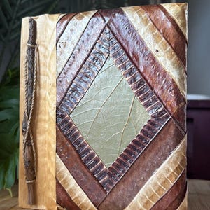 May include: A rectangular journal with a light wood spine and a cover featuring a geometric pattern of layered, dried leaves in shades of brown, tan, and pale green. A thin, dark twig is attached to the spine with a light-colored cord.