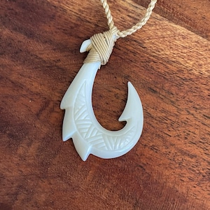 May include: A white, carved bone pendant in the shape of a fish hook, with a detailed geometric pattern. The hook is suspended from a beige, braided cord. The cord is attached to the hook with a wrapped, beige twine.