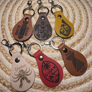 May include: A collection of six leather keychains in various colors, each with a silver keyring and clasp. The keychains feature engraved designs and text, including a bomb, Guam seal, palm tree, crab, and the words "Guam," "Guahan," and "Hafa Adai."