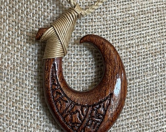May include: A handcrafted wooden pendant necklace featuring a dark brown, curved hook design. The pendant has intricate carved details and is suspended from a natural-colored cord. The necklace is displayed on a textured, neutral-toned surface.