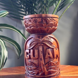 May include: A brown wooden carved cup featuring a relief design of a palm tree and the text GUAM. The cylindrical vessel has a wide, flared rim and a base with decorative carved patterns, showcasing the natural wood grain and texture throughout.