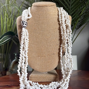 May include: A multi-strand necklace composed of numerous small, white, textured seashells. The strands are gathered together and secured by a single, larger, conical-shaped shell bead featuring a distinct black and white patterned design.