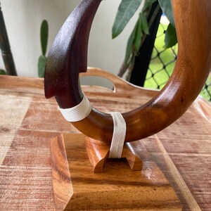 May include: A handcrafted wooden hook sculpture with a curved design. The hook is made of dark brown wood with a lighter brown section and is secured with a white woven band. It is mounted on a wooden base.
