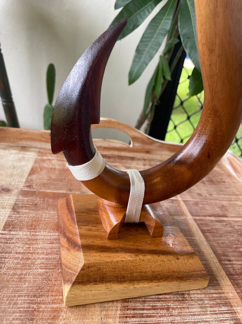 May include: A handcrafted wooden hook sculpture with a curved design. The hook is made of dark brown wood with a lighter brown section and is secured with a white woven band. It is mounted on a wooden base.