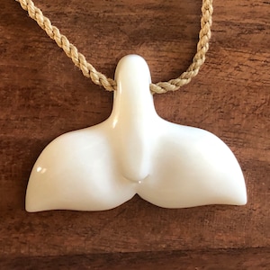 Puede incluir: Collar con colgante de cola de ballena blanca. El colgante blanco brillante cuelga de un cordón trenzado beige. El collar se exhibe sobre una superficie de madera.