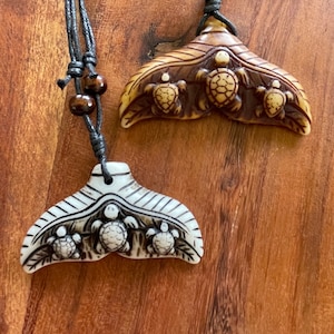 Op de afbeelding: Twee hangerkettingen met een walvisstaartontwerp, elk met drie gebeeldhouwde zeeschildpadden. Eén hanger is gebroken wit, de andere is bruin. Beide zijn geregen aan zwarte koorden met houten kralen, tegen een houten achtergrond.