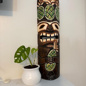Puede incluir: Una máscara tiki alta de madera marrón oscuro con detalles en verde y blanco. La máscara presenta diseños de piña tallados y una cara detallada. Una pequeña maceta blanca con una planta verde se encuentra junto a la máscara.
