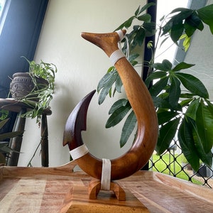 May include: A wooden sculpture of a fish hook, with a dark brown hook and a lighter brown curved body. The word "HAWAII" is carved into the top. White cord wraps around the hook and body. The sculpture sits on a wooden base.