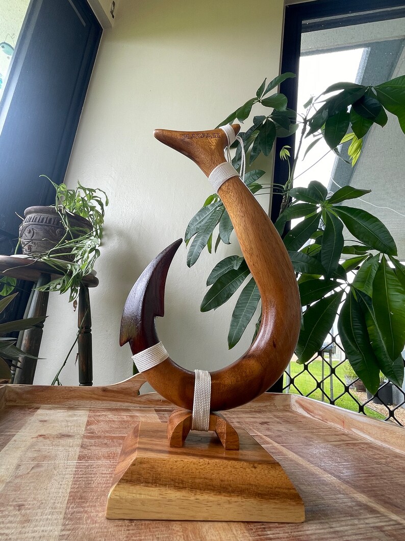 May include: A wooden sculpture of a fish hook, with a dark brown hook and a lighter brown curved body. The word "HAWAII" is carved into the top. White cord wraps around the hook and body. The sculpture sits on a wooden base.