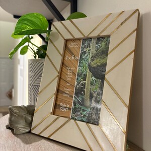 Op de afbeelding: Crèmekleurige fotolijst met gouden diagonale accenten. De lijst bevat een foto van 10 x 15 cm en is gemarkeerd met "Made in GUAM". De lijst is gemaakt met botinlegwerk en kokospalm-achterkant. Een decoratief item voor woondecoratie.
