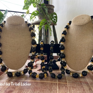 May include: Several necklaces on display. Each necklace features large, glossy black beads interspersed with smaller white and yellow beads. The necklaces are displayed on tan busts, with the text "Pasefika Tribal Locker" visible.