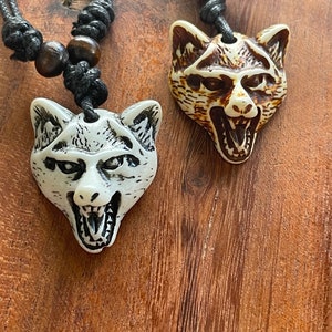 May include: Two wolf head pendants on black cords with wooden beads. The pendants are carved with detailed features, one in off-white, the other in brown and cream. The pendants are displayed on a wooden surface.